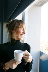 Happy young woman at home drinking cup of coffee looking out of window - VABF01138