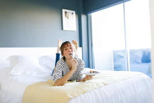 Young woman eating piece of vegan cake, lying on bed, licking spoon - VABF01199