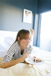 Junge Frau isst ein Stück veganen Kuchen, liegt auf dem Bett und leckt den Löffel - VABF01200