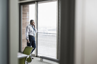 Ein leitender Angestellter im Büro steht am Fenster und hält ein Smartphone in der Hand - UUF09920