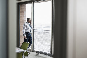 Senior manager in office standing at the window holding smartphone - UUF09920