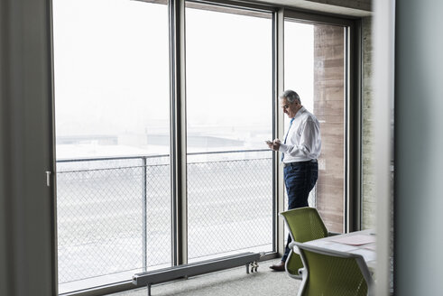 Ein leitender Angestellter im Büro steht am Fenster und hält ein Smartphone in der Hand - UUF09921