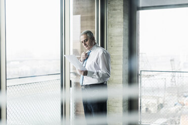 Senior manager in office standing at window talking on smartphone, holding document - UUF09937