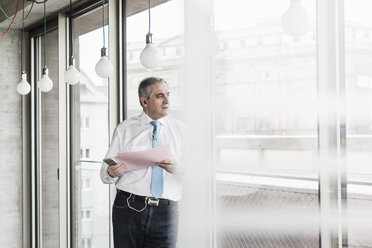 Senior manager in office standing at window talking on smartphone, holding document - UUF09938