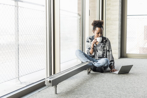 Junge Frau sitzt auf dem Boden, benutzt einen Laptop und trinkt Kaffee - UUF09952