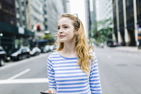 USA, New York, Manhattan, Young woman walking in the street, holding mobile phone - GIOF01891