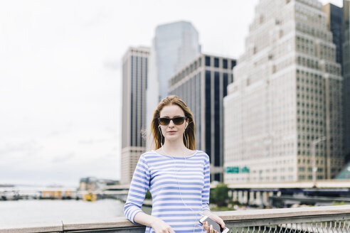 USA, New York City, Junge Frau steht in Manhattan an einem Geländer und hält ein Smartphone in der Hand - GIOF01910