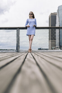 USA, New York City, Junge Frau steht in Manhattan an einem Geländer und hält ein Smartphone in der Hand - GIOF01913