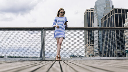 USA, New York City, Junge Frau steht in Manhattan an einem Geländer und hält ein Smartphone in der Hand - GIOF01914