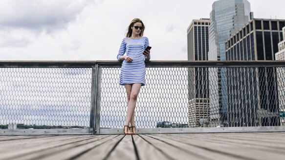 USA, New York City, Junge Frau steht in Manhattan an einem Geländer und hält ein Smartphone in der Hand - GIOF01914