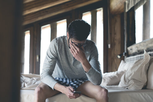 Tired young man with smartphone sitting on bed at home squinting - ZOCF00142