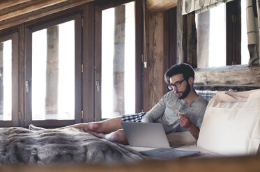 Junger Mann mit Laptop und Kreditkarte zu Hause im Bett liegend - ZOCF00144