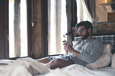 Young man using smartphone lying in bed at home - ZOCF00147