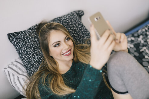 Lächelnde junge Frau liegt auf dem Bett und benutzt ein Mobiltelefon - LCUF00090