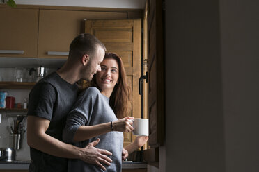 Amorous couple standing in kitchen, embracing with cup of coffee - KKAF00451