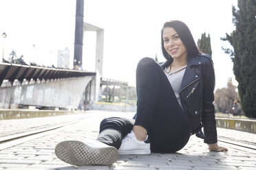 Portrait of smiling young woman wearing black leather jacket sitting on pavement - ABZF01934