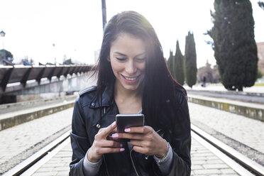 Porträt einer lächelnden jungen Frau in schwarzer Lederjacke, die auf ihr Handy schaut - ABZF01935