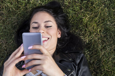 Porträt einer lächelnden jungen Frau mit Nasenpiercing, die auf einer Wiese liegt und auf ihr Handy schaut - ABZF01936