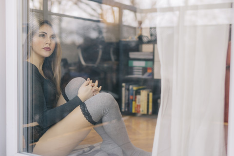 Junge Frau sitzt auf dem Boden des Wohnzimmers und schaut durch die Balkontür, lizenzfreies Stockfoto