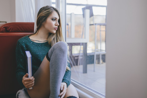 Junge Frau mit Buch sitzt auf dem Boden des Wohnzimmers und schaut durch die Balkontür, lizenzfreies Stockfoto