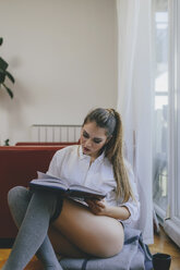 Junge Frau sitzt auf dem Boden des Wohnzimmers und liest ein Buch - LCUF00098