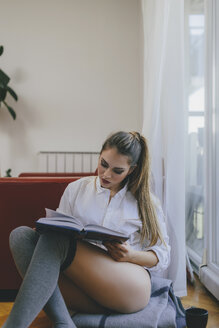 Young woman sitting on the floor of living room reading a book - LCUF00098