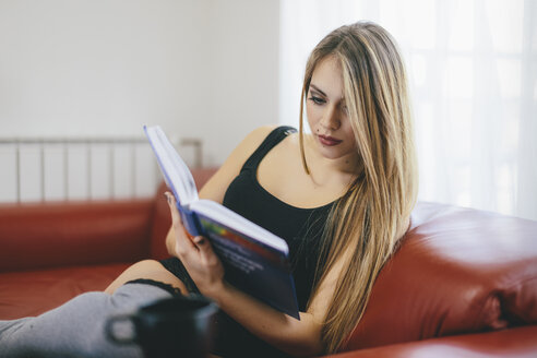 Junge Frau sitzt auf der Couch und liest ein Buch - LCUF00099