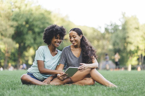 Zwei beste Freunde sitzen auf einer Wiese in einem Park und haben Spaß mit einem Mini-Tablet, lizenzfreies Stockfoto