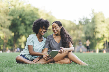 Two best friends sitting on a meadow of a park having fun with mini tablet - GIOF01993