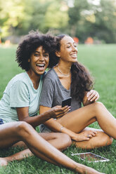 Two best friends sitting on a meadow of a park having fun with mini tablet - GIOF01994