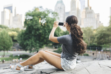 USA, Manhattan, junge Frau sitzt im Central Park und fotografiert die Skyline mit ihrem Smartphone - GIOF01999