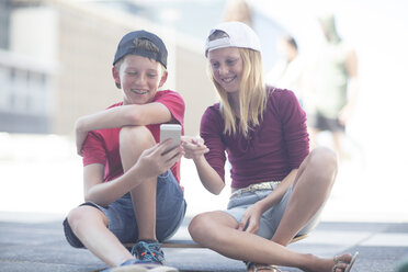 Junge und Mädchen benutzen ein Smartphone, sitzen auf einem Skateboard - ZEF12902