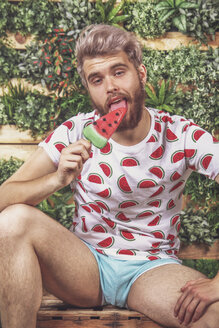 Porträt eines jungen Mannes, der auf der Terrasse vor einem vertikalen Garten einen Wassermelonen-Eislutscher isst - RTBF00692