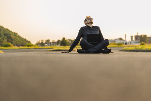 Businessman sitting on rural road with laptop - KNSF01055