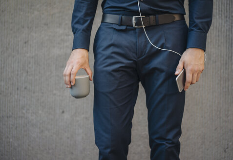 Businessman holding smartphone with connected earphones and cup of coffee - KNSF01064