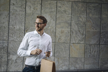 Smiling businessman holding cell phone and paper bag - KNSF01067