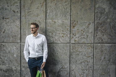 Smiling man holding paper bags standing at a wall - KNSF01068