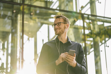 Smiling businessman using smartphone with connected earphones - KNSF01118