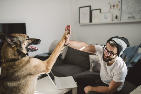 Junger Mann zu Hause gibt High Five mit seinem Hund, lizenzfreies Stockfoto