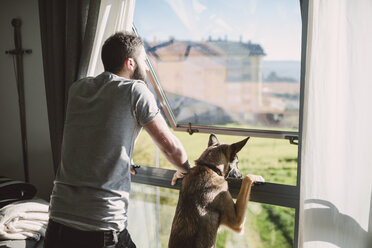 Young man standing at window with his dog, waiting - RAEF01748