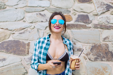 Junge Frau am Strand mit einer Tasse Kaffee und einem Smartphone, lizenzfreies Stockfoto