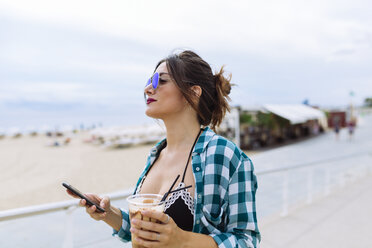 Junge Frau am Strand mit einer Tasse Kaffee und einem Smartphone - GIOF02011