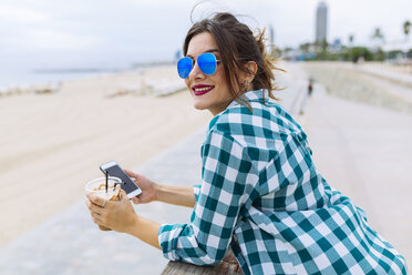 Junge Frau am Strand mit einer Tasse Kaffee und einem Smartphone - GIOF02012