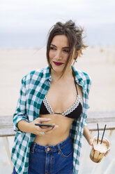 Junge Frau am Strand mit einer Tasse Kaffee und einem Smartphone - GIOF02013