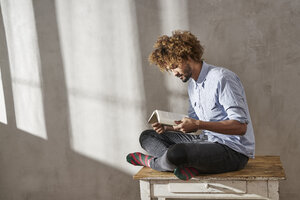 Junger Mann sitzt auf einem Holztisch und liest ein Buch - FMKF03506