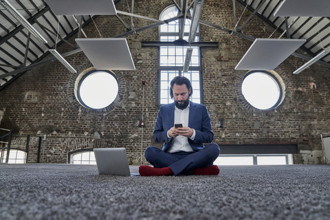 Geschäftsmann sitzt auf Teppich in einem Loft und benutzt ein Mobiltelefon, lizenzfreies Stockfoto