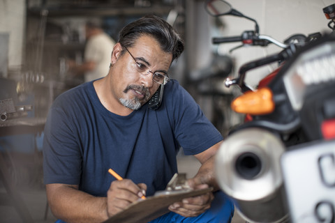 Mechaniker mit Klemmbrett und Telefon in einer Motorradwerkstatt, lizenzfreies Stockfoto