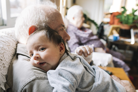 Urgroßvater hält Baby zu Hause mit seiner Frau im Hintergrund, lizenzfreies Stockfoto