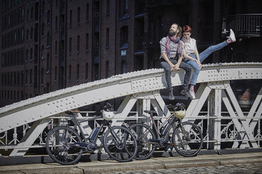 Deutschland, Hamburg, Paar mit Elektrofahrrädern auf Brückengeländer sitzend - RORF00649