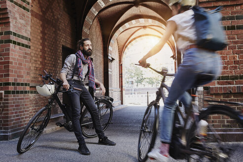 Man with electric bicycle watching woman passing by on bicycle - RORF00671